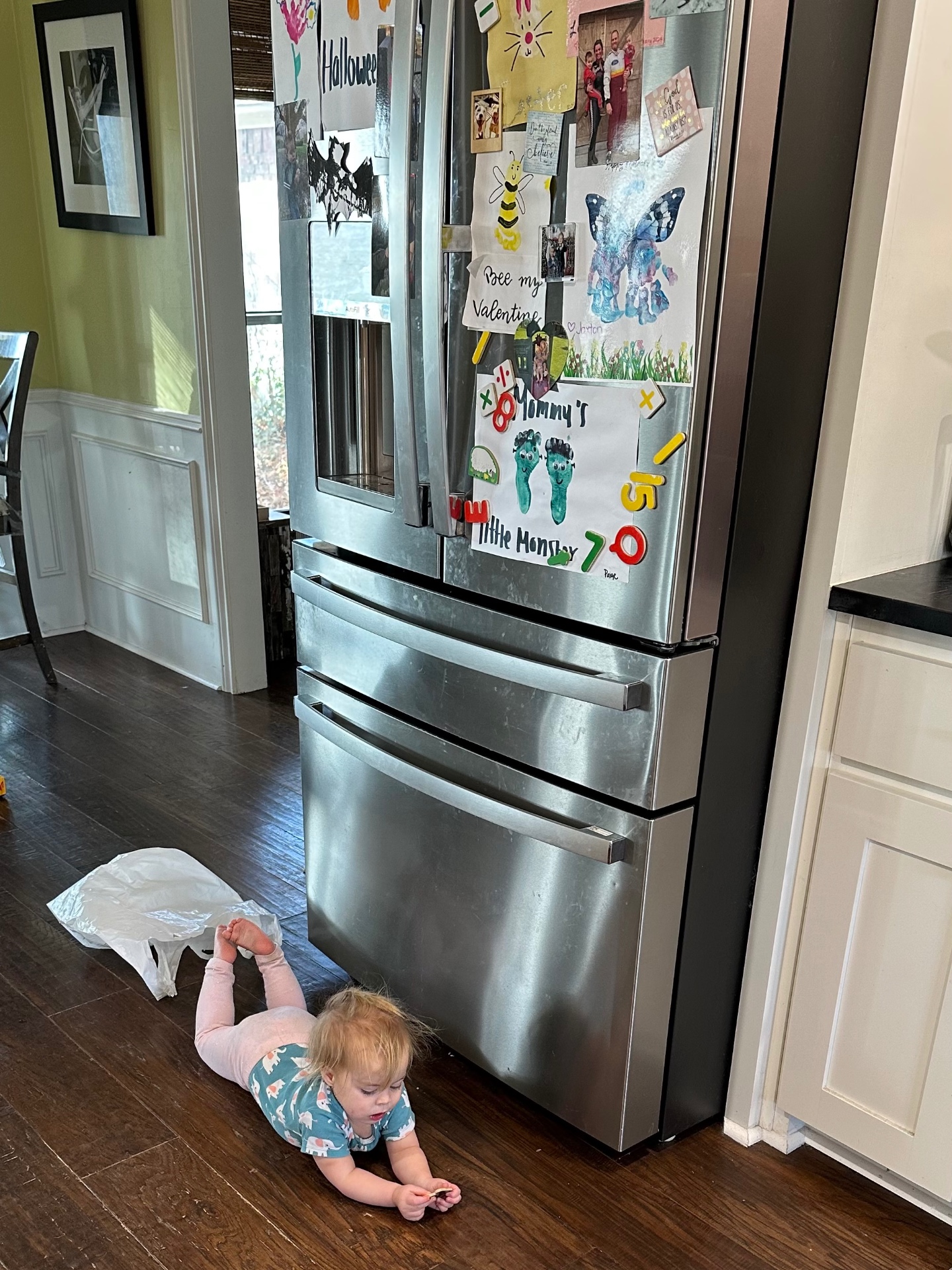 Family refrigerator covered in the kids' artwork and magnetic letters, with the baby playing on the kitchen floor